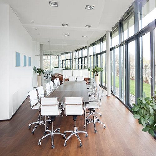 Spacious and bright business conference room with hard wood floor and a bank of windows, opening up into a garden. There are chairs lines up and the decoration is very modern and contemporary.